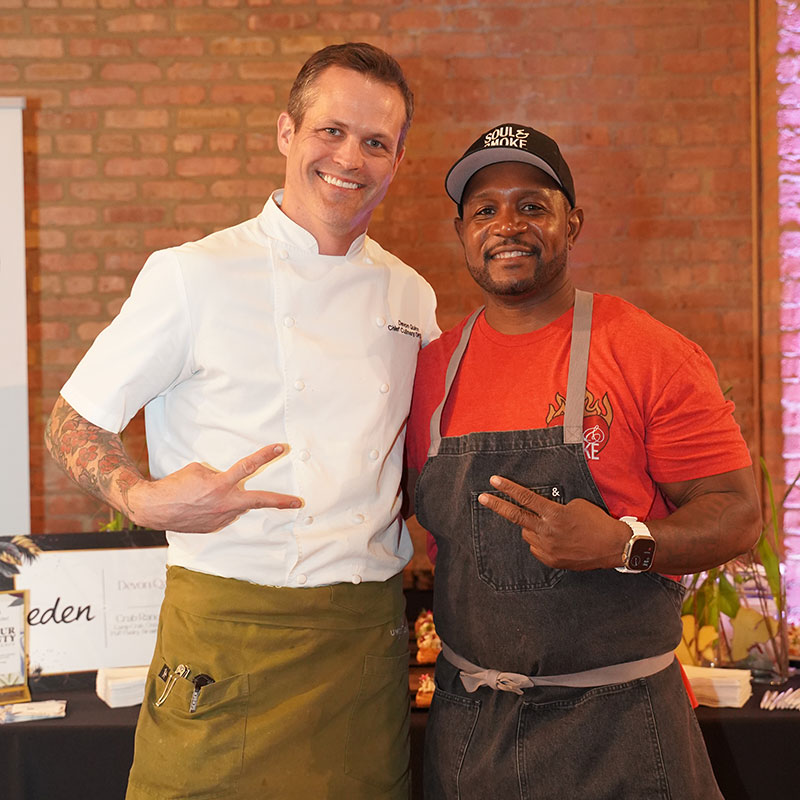 Chef Devon Quinn of Paramount Events and Chef D’Andre Carter of Soul & Smoke stand side by side smiling, Chef Quinn is in a white chef’s coat and green apron, Chef D'Andre Carter in a red shirt, gray apron, and black cap. Both gesture with peace signs. Exposed brick wall background.