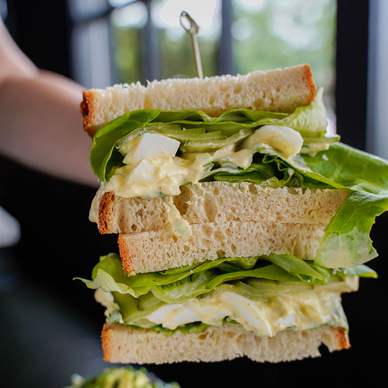 Close-up of eden's august sandwich collab, a halved deviled egg salad sandwich with thick slices of bread, leafy lettuce, and visible chunks of boiled egg, held together by a skewer, with a blurred background.