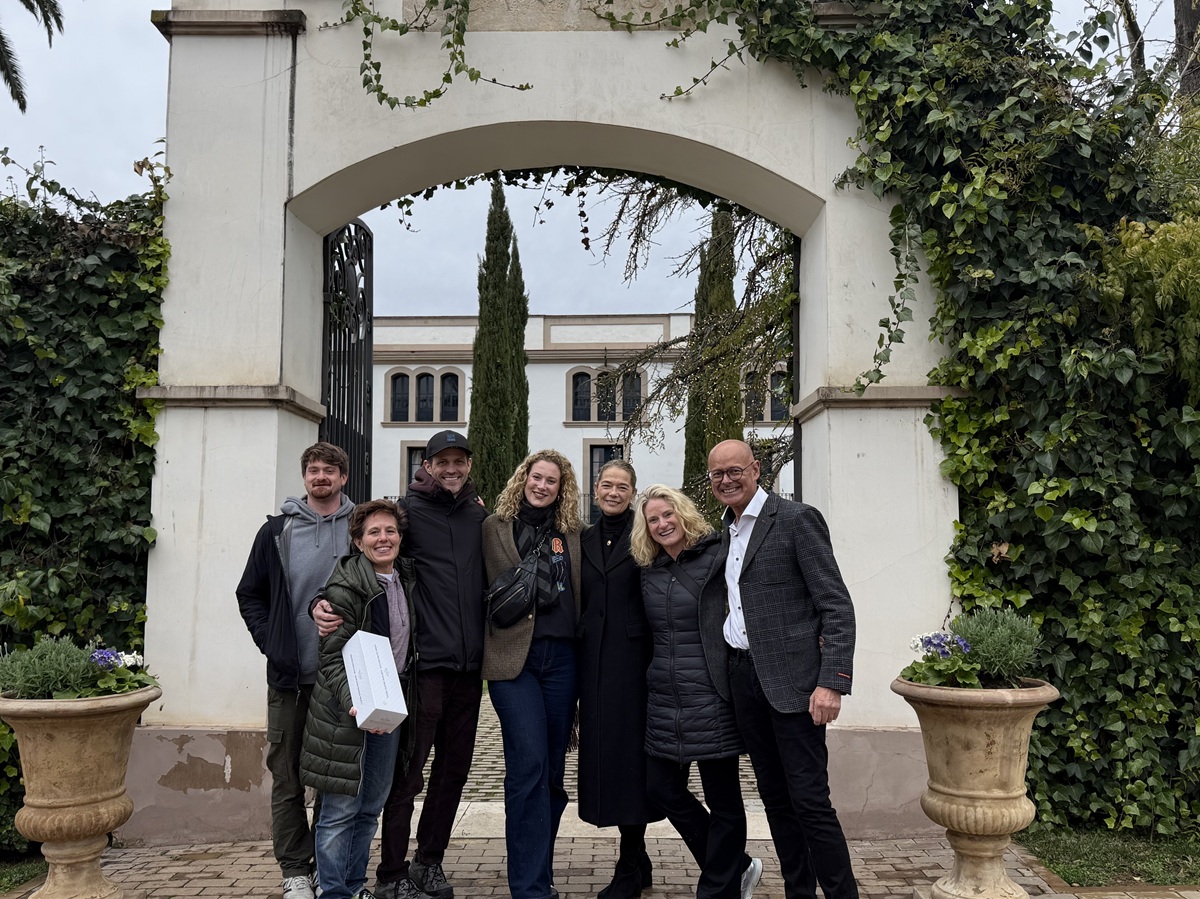 Six people stand smiling together under a large white archway covered in ivy, with a building and tall trees in the background. Two large planters with flowers frame the scene, as one person holds a sandwich, adding a casual touch to the moment.