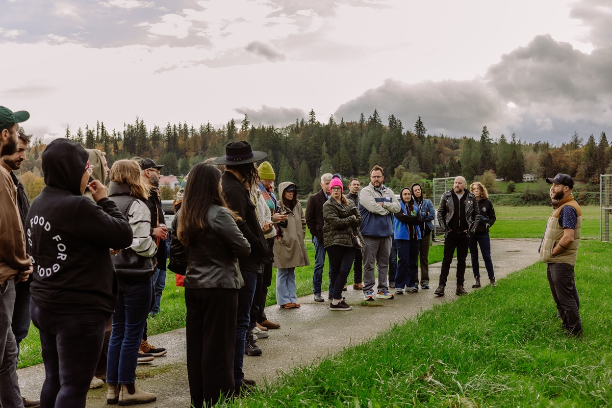 A group of people stands outdoors on a path in a grassy field, listening to a person speaking. Trees and cloudy skies are visible in the background. Most people are dressed warmly in jackets and hats to honor chef devon quinn of eden