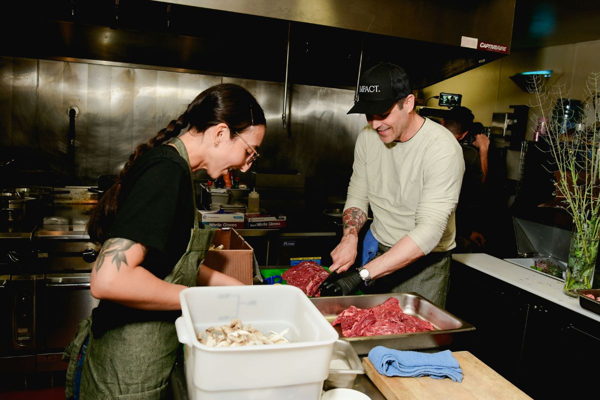 chef devon quinn with another chef in a commercial kitchen smile as they prepare food; one, possibly chef Devon Quinn, holds a tray of raw meat, while the other sorts mushrooms into a white container. Stainless steel surfaces and kitchen equipment gleam in the background.
