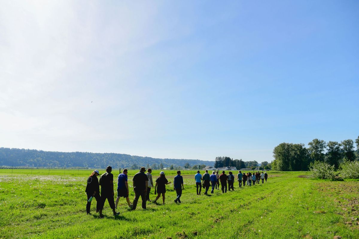 A group of people, led by Devon Quinn, walk in a line across a grassy field under a clear blue sky, with trees and hills visible in the background.