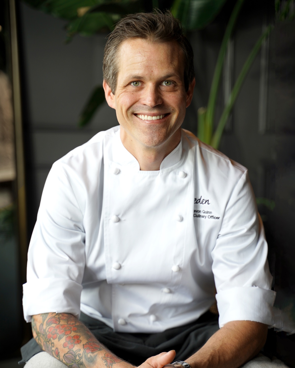 Chef Devon Quinn of eden smiles at the camera in a white chefs coat sits with his hands in his lap. He has tattoos on one forearm and is in front of a dark background with green plants visible.