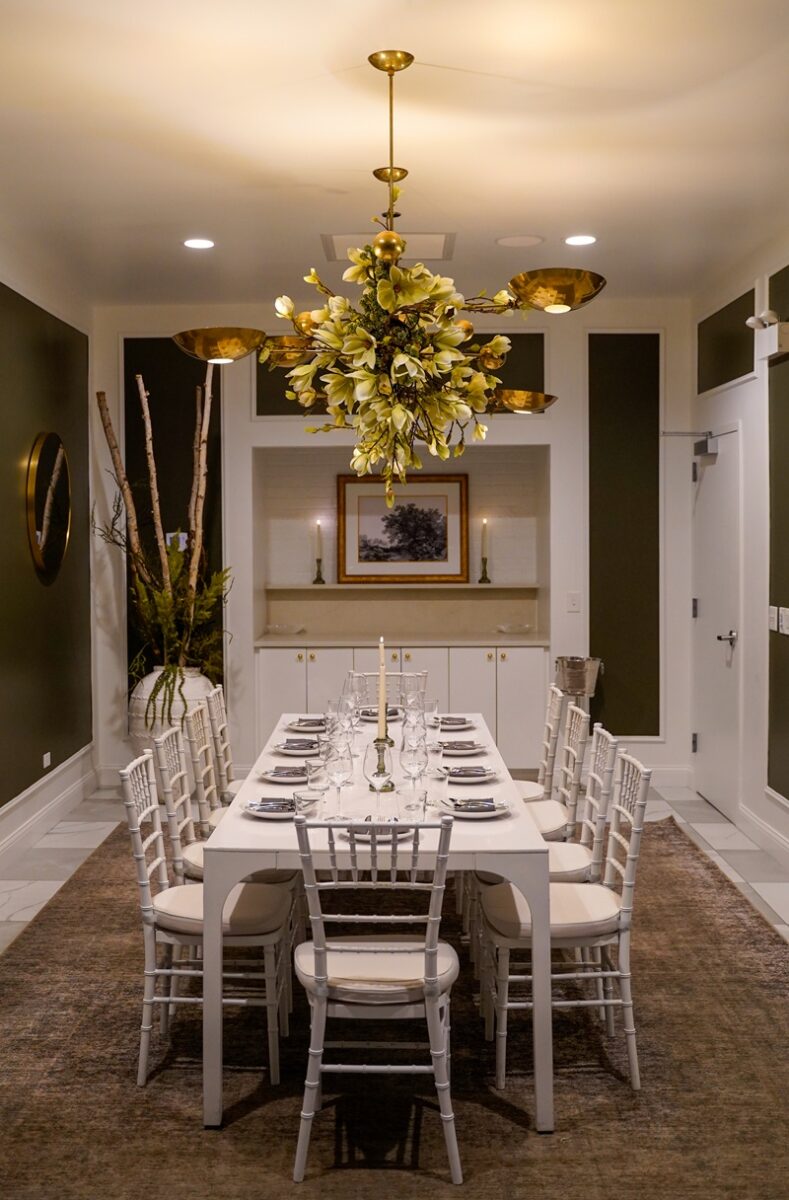 A modern private dining room with a white table set for eight, white chairs, a golden floral chandelier, candles, wall art, and green walls with white trim creates an elegant, inviting atmosphere.