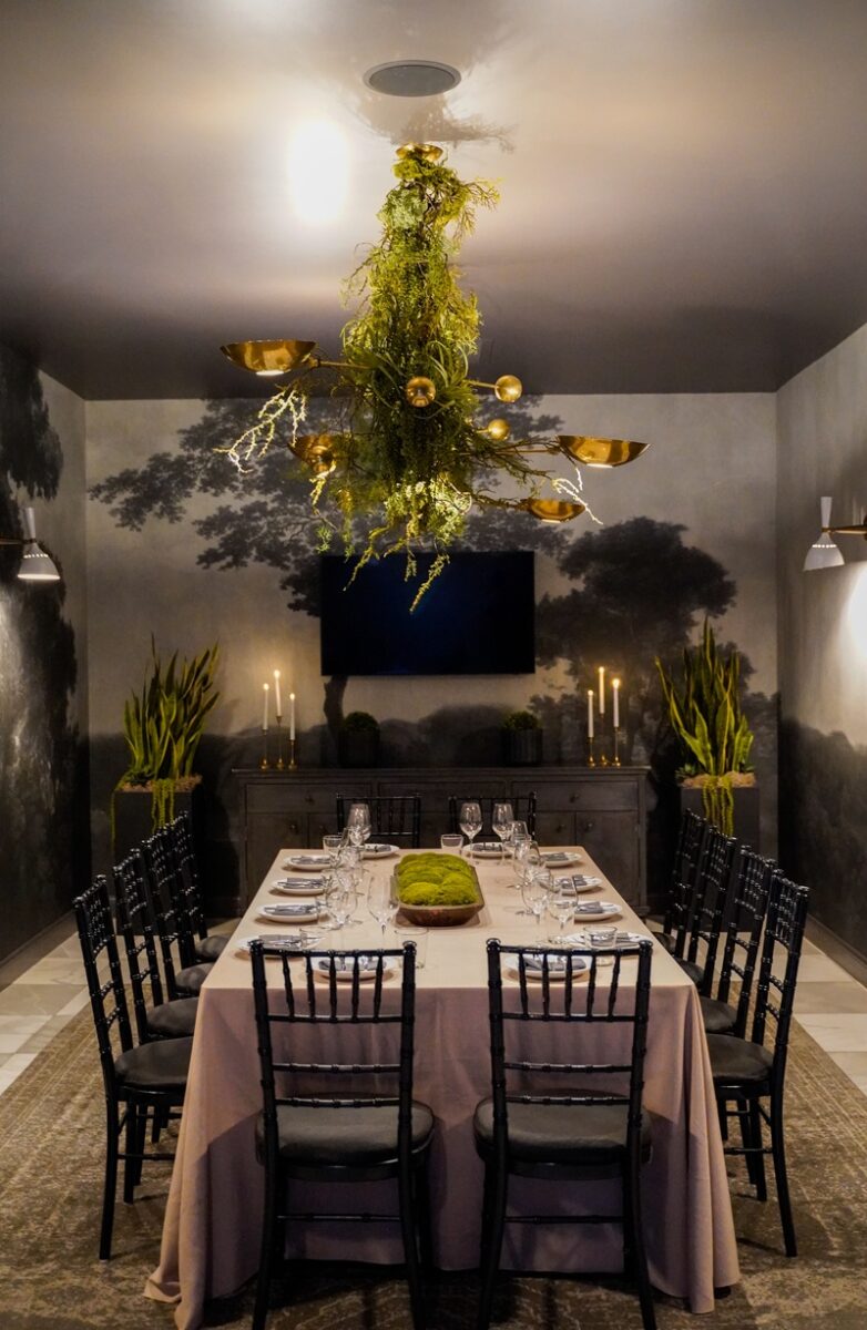 A formal private dining room with a rectangular table set for twelve, black chairs, a moss centerpiece, greenery on the chandelier, wall candles, potted plants, and nature-themed wall art in a dim, elegant setting.