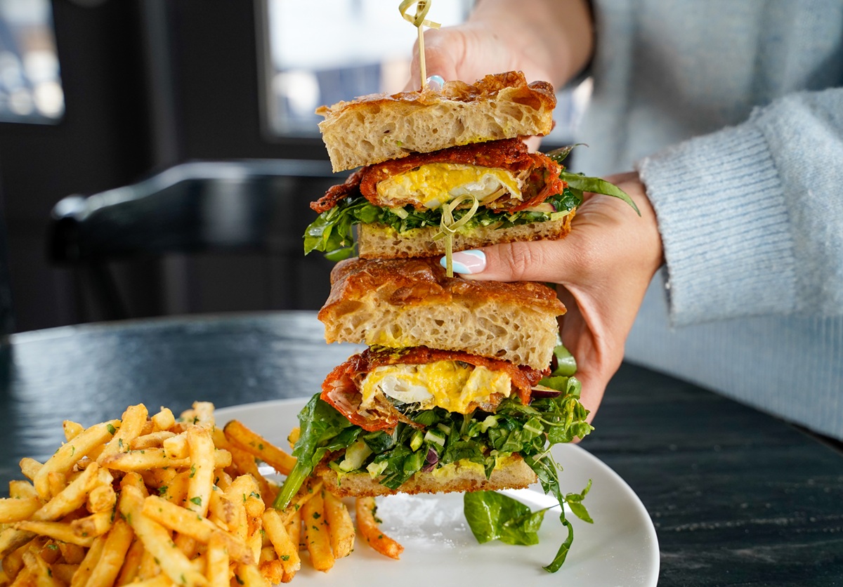 A person holds a large stacked sandwich filled with greens, egg, and meat between thick slices of bread, with a plate of seasoned French fries next to it on a dark table.