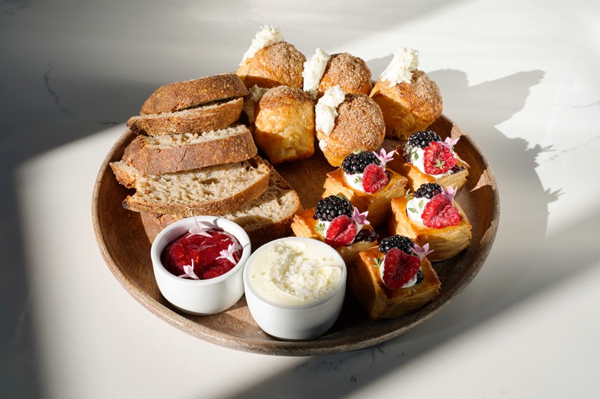 A wooden plate with sliced bread, cream-filled pastries, fruit-topped danishes, and two small bowls containing jam and whipped butter or cream, from eden's bakery program in chicago