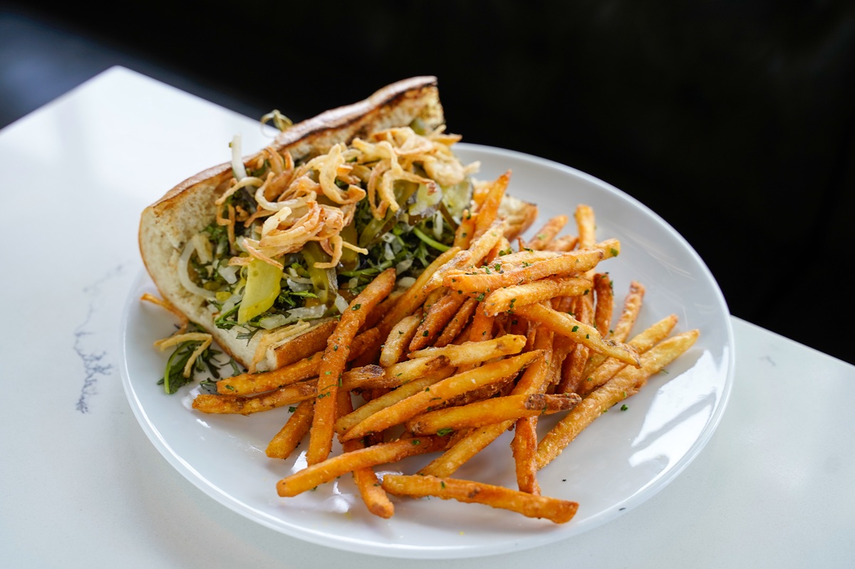 A white plate holds a delicious sandwich packed with greens and crispy onions, served with a generous portion of seasoned French fries on a white table.