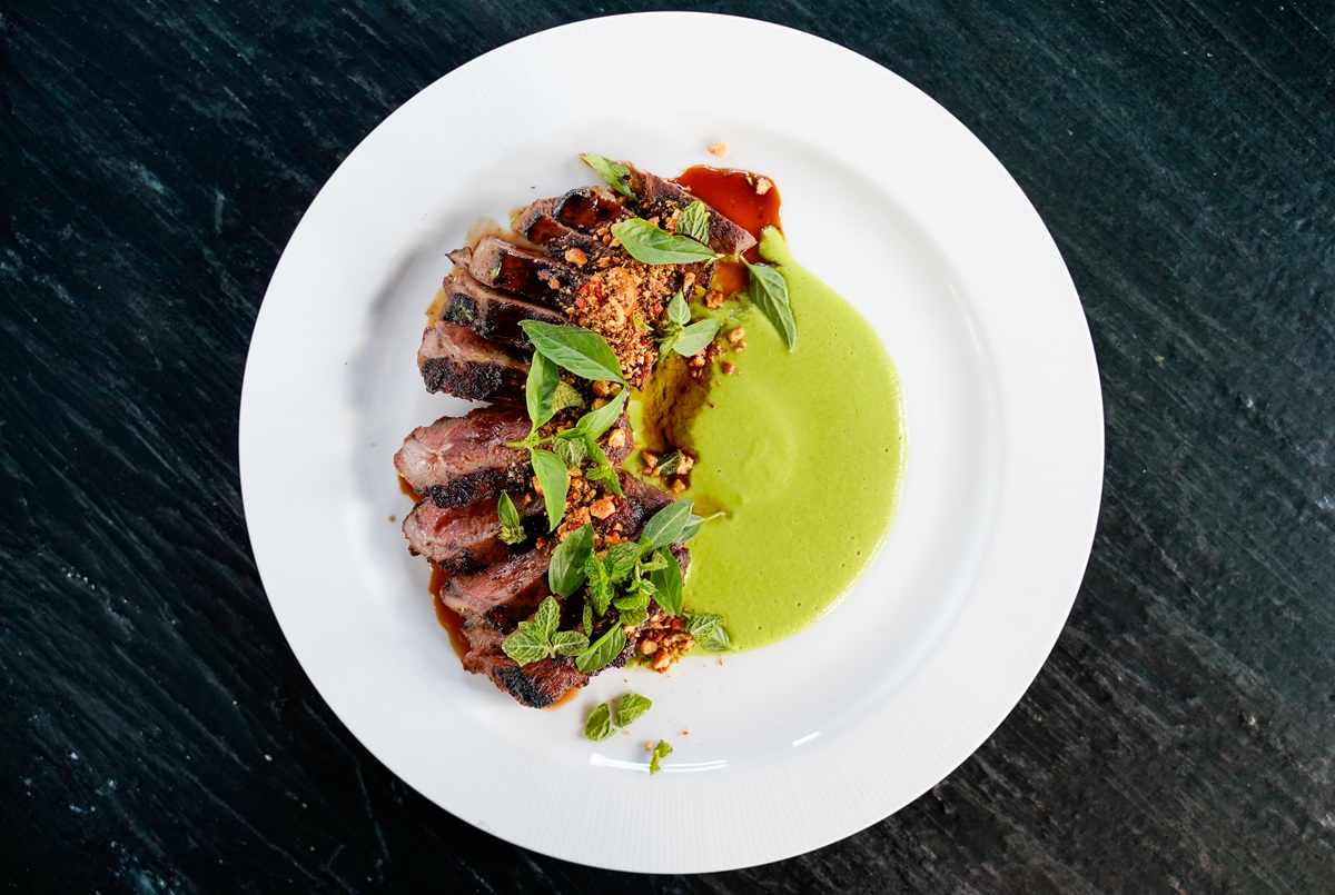 Sliced grilled steak served on a white plate with green sauce, garnished with fresh herbs and crushed nuts, placed on a dark textured surface.