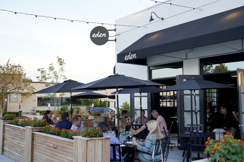 People dining outdoors under black umbrellas at a restaurant called Eden, with flower boxes along a wooden patio fence and a black awning above the entrance on a clear day.