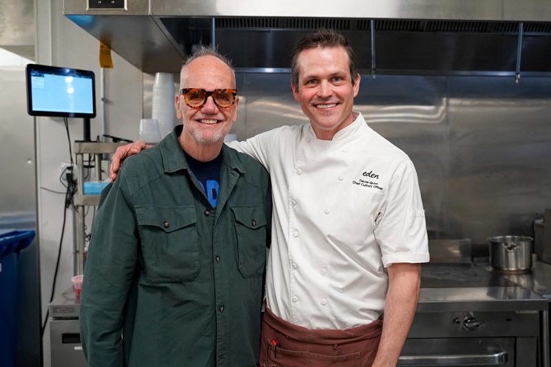 Chef Devon Quinn and Chef Tony Mantuano stand in a kitchen, ready to craft the perfect sandwich. One wears sunglasses and a green jacket, while the other sports a white chef’s coat and brown apron. Stainless steel appliances and cooking equipment are visible in the background.