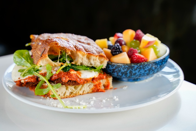 A delicious eggplant parmasean sandwich for eden's september chicago sandwich collab with greens and tomato sauce sits on a white plate next to a blue bowl filled with assorted fresh fruit, including berries, grapes, and apple pieces.